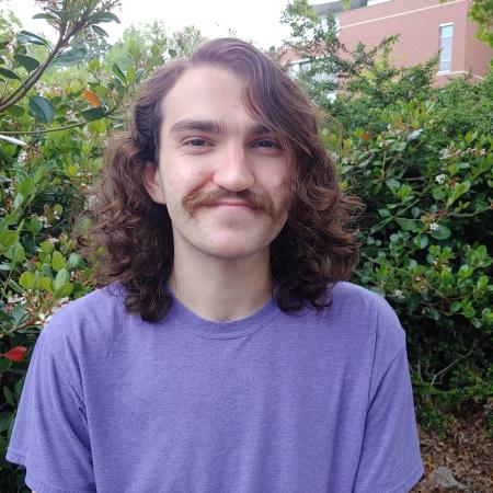 Ian smiling while sitting on a bench outside. A green shrub covers most of the background.